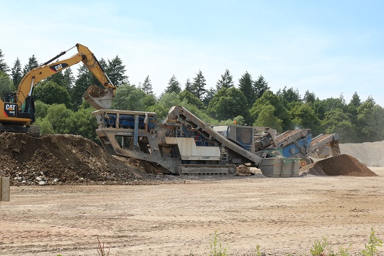 Recycling Bereich Bagger im Hintergrund. Im Vordergrung ein Fläche mit Sand.